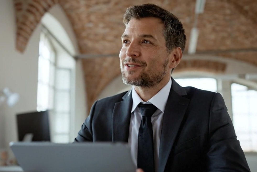 Ein professioneller, sympathischer Immobilienmakler im dunklen Anzug sitzt mit seinem Laptop in einem stilvollen Büro mit historischer Backstein-Gewölbedecke. Er blickt aufmerksam und freundlich auf, passend zu einem positiven Beratungsgespräch mit Kunden.